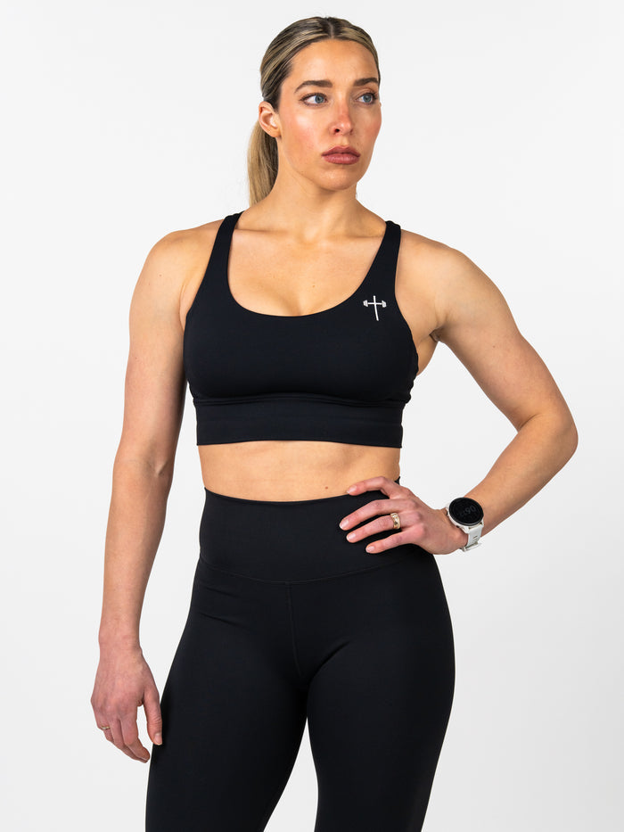Woman wearing a black Essential Sports Bra featuring a small Christian cross and barbell logo on the chest, paired with high-waisted black leggings in a fitness studio setting.