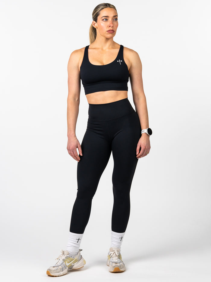 Woman wearing a black Essential Sports Bra with a small cross logo on the chest, paired with black leggings and white socks featuring a cross symbol, styled for Christian fitness apparel.