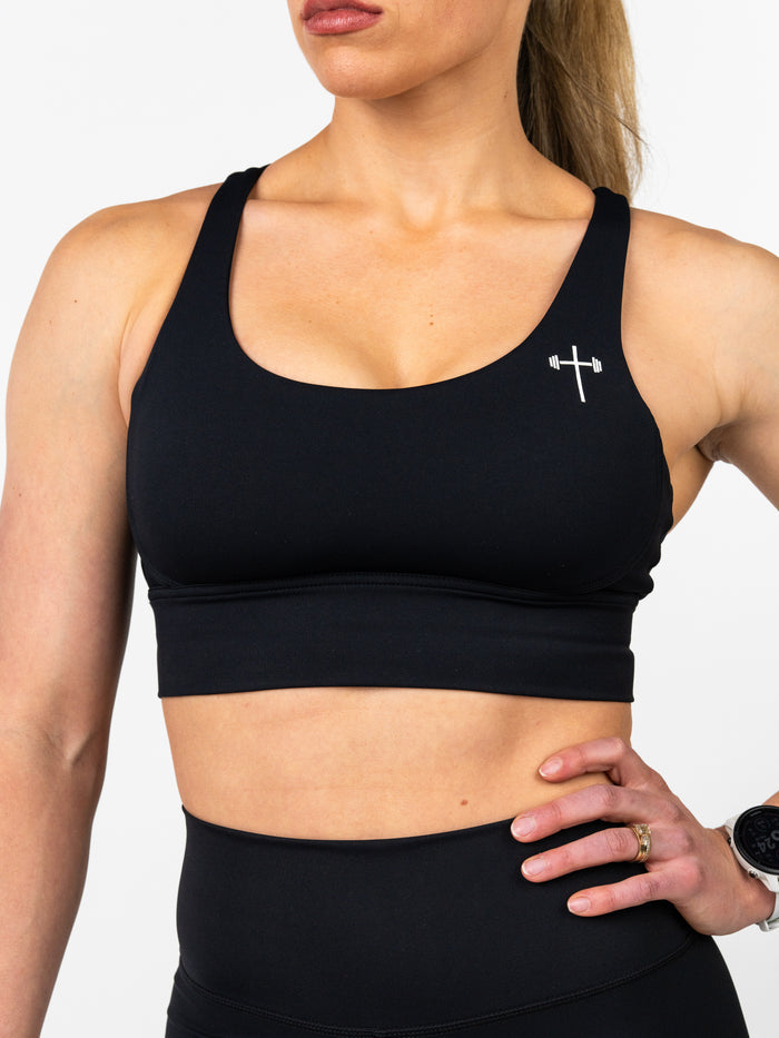 Woman wearing a black Essential Sports Bra featuring a small white Christian cross with a barbell logo on the chest, designed for high performance and extreme comfort.