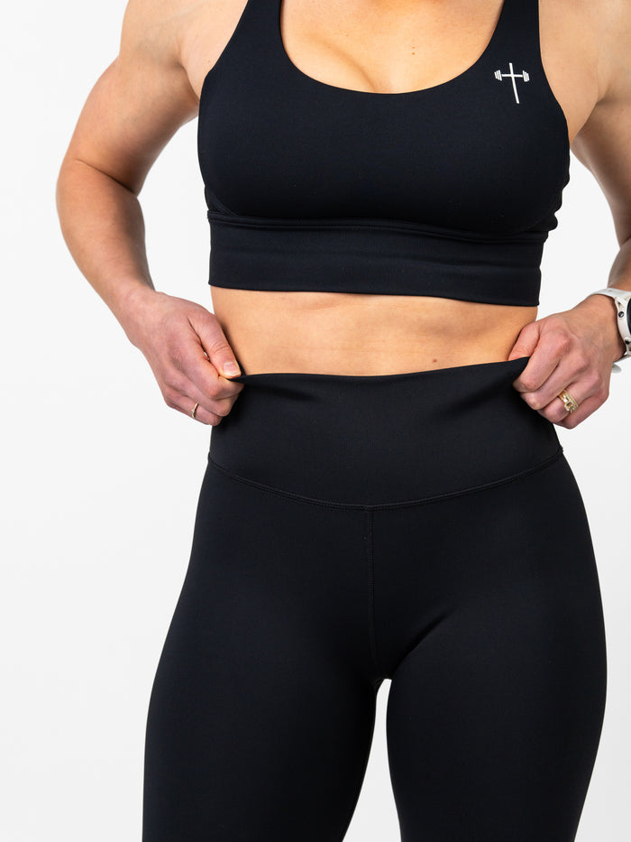 Woman wearing a black Essential Sports Bra featuring a small cross and barbell logo, paired with high-waisted black leggings, showcasing flexible and comfortable Christian fitness apparel.