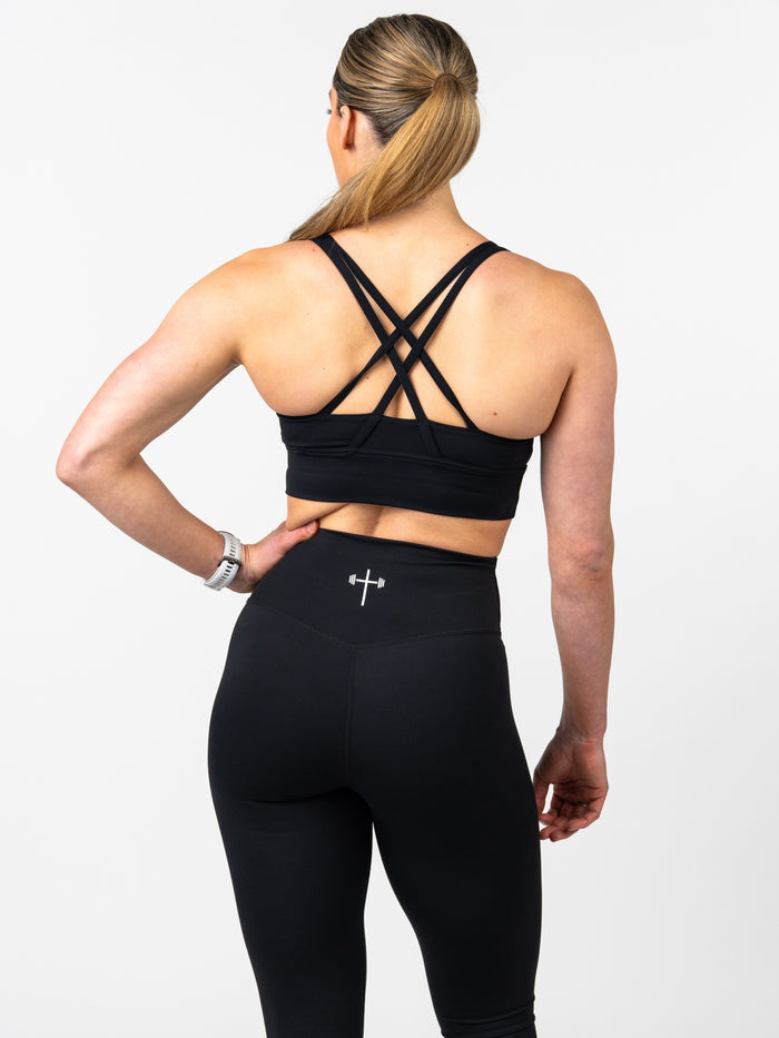 Rear view of a woman wearing the Essential Sports Bra with a unique crisscross strap design and matching black leggings featuring a small Christian cross with barbell logo, showcasing faith based fitness apparel.
