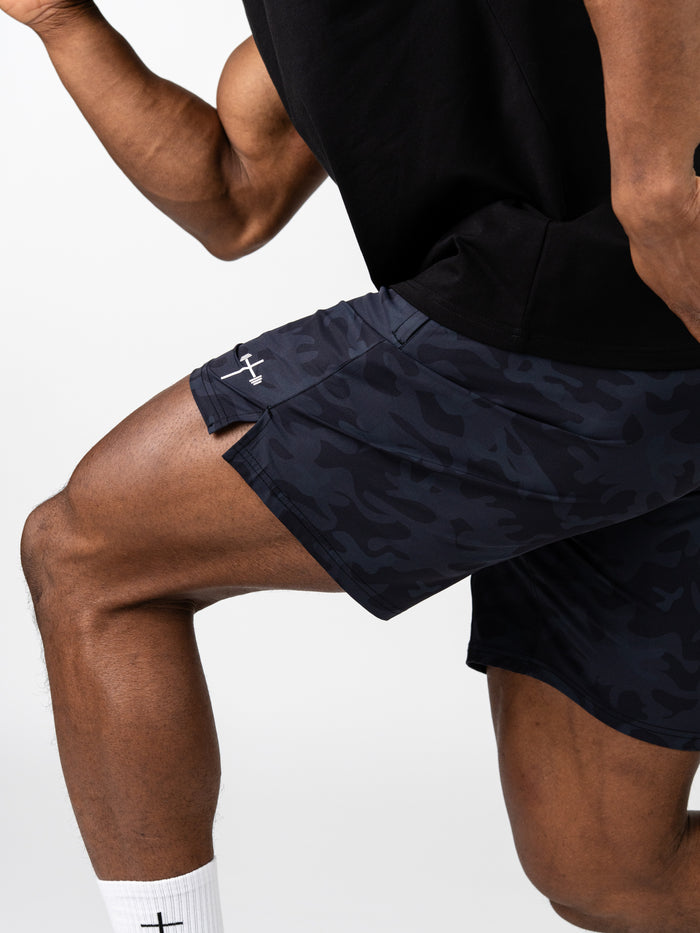 Close-up of a person performing a lunge wearing black camo Essential Training Shorts with a small white cross and barbell symbol on the side, paired with a black shirt and white socks.