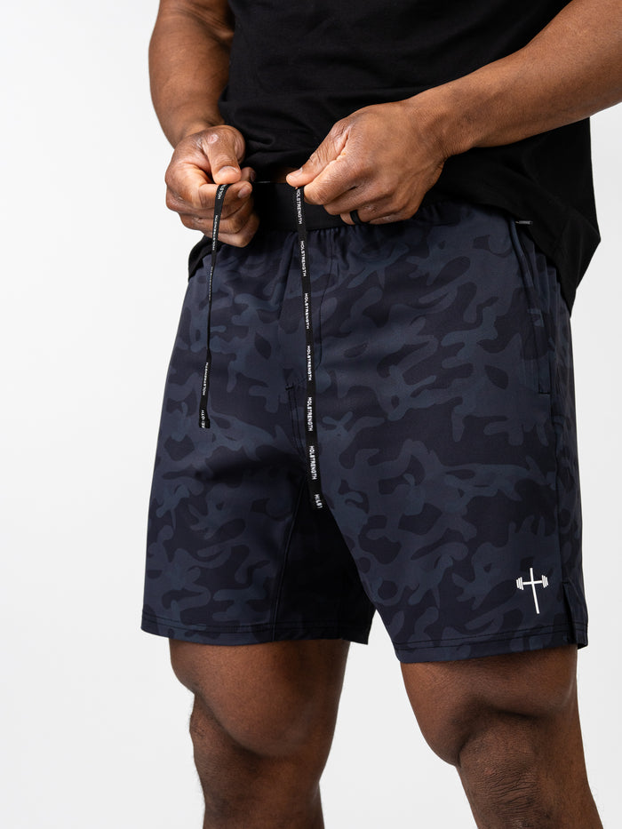 Man adjusting black camo Essential Training Shorts with drawstring and discreet logo on left leg, designed for comfort and movement during workouts.