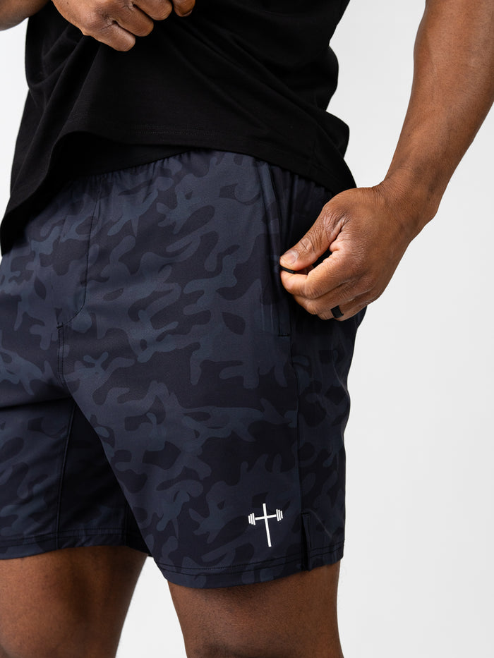 Close-up of a person wearing Essential Training Shorts in dark camo pattern with a subtle white barbell cross logo near the hem, highlighting lightweight, breathable material.
