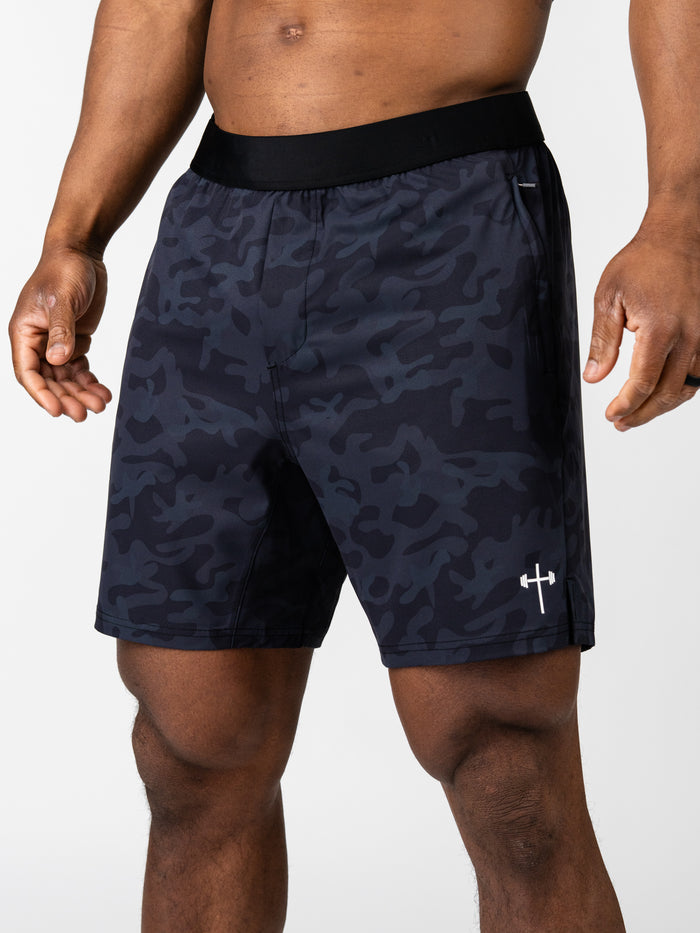 Close-up of a muscular man wearing Essential Training Shorts in dark camo with a subtle cross and barbell logo near the hem, designed for faith-based fitness activities.