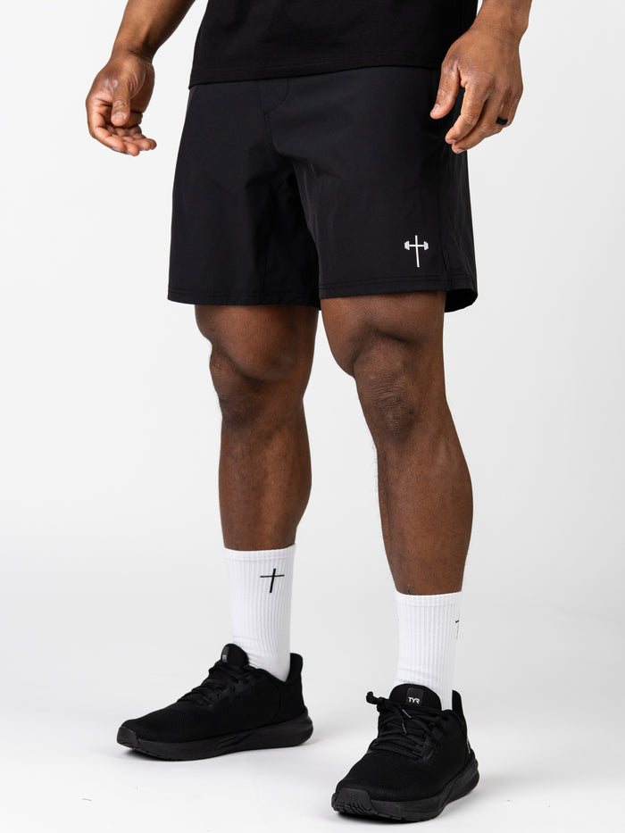 A man wearing black Essential Training Shorts featuring a small white cross and barbell logo on the left leg, paired with white crew socks with cross embroidery and black athletic shoes.