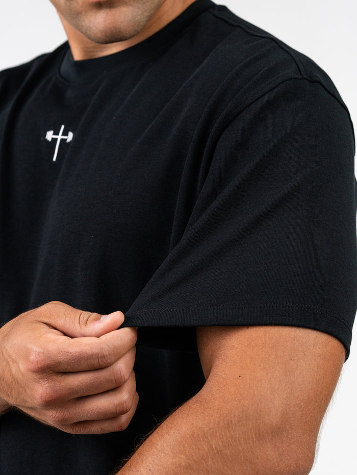 Close-up of a person wearing the Faith Over Fear Premium Oversized Tee in black, showcasing its heavyweight fabric and relaxed fit with a small white barbell cross logo on the chest.
