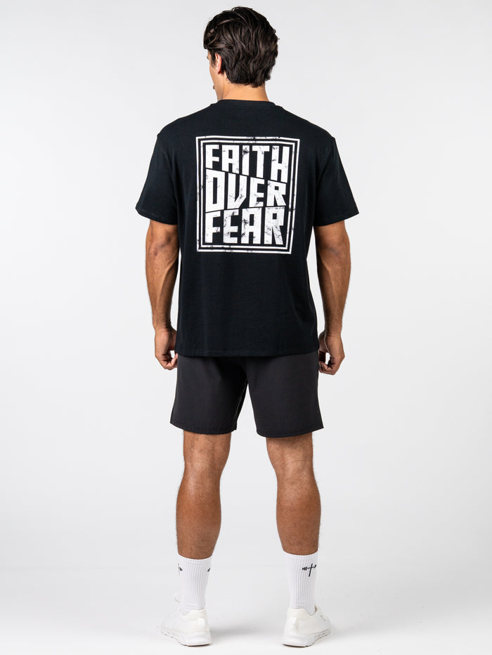 Man wearing the Faith Over Fear Premium Oversized Tee in black with bold white text on the back, paired with black shorts and white athletic shoes, showcasing Christian fitness apparel.