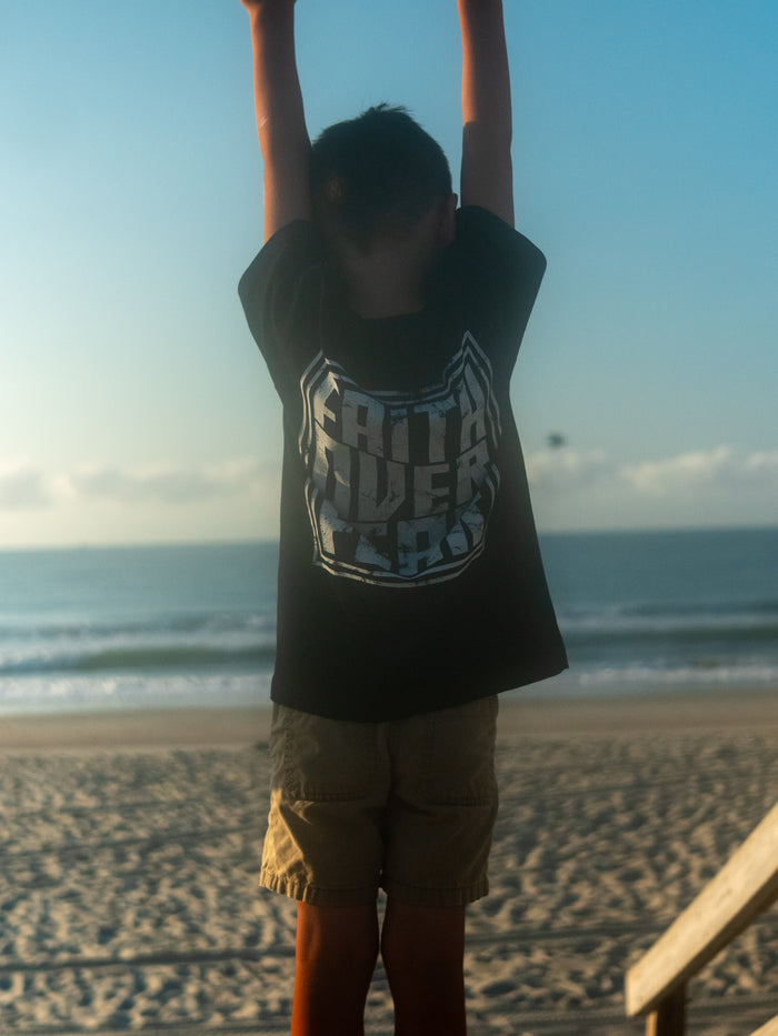 Child wearing a Faith Over Fear Youth Tee with an oversized graphic on the back, standing with arms raised at a beach during sunrise or sunset.