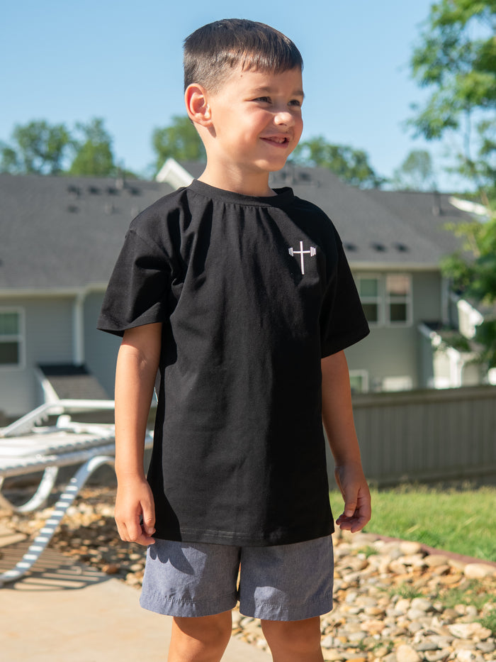 Young boy standing outdoors wearing a black Jesus is King Youth Tee featuring a small white cross logo on the front, paired with gray shorts on a sunny day.