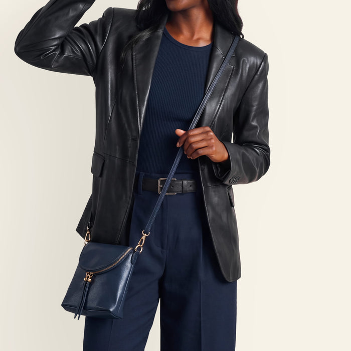 A model wearing a black leather blazer, navy blue ribbed top, navy trousers, and carrying a navy blue leather crossbody bag with gold-tone hardware against a light beige background.