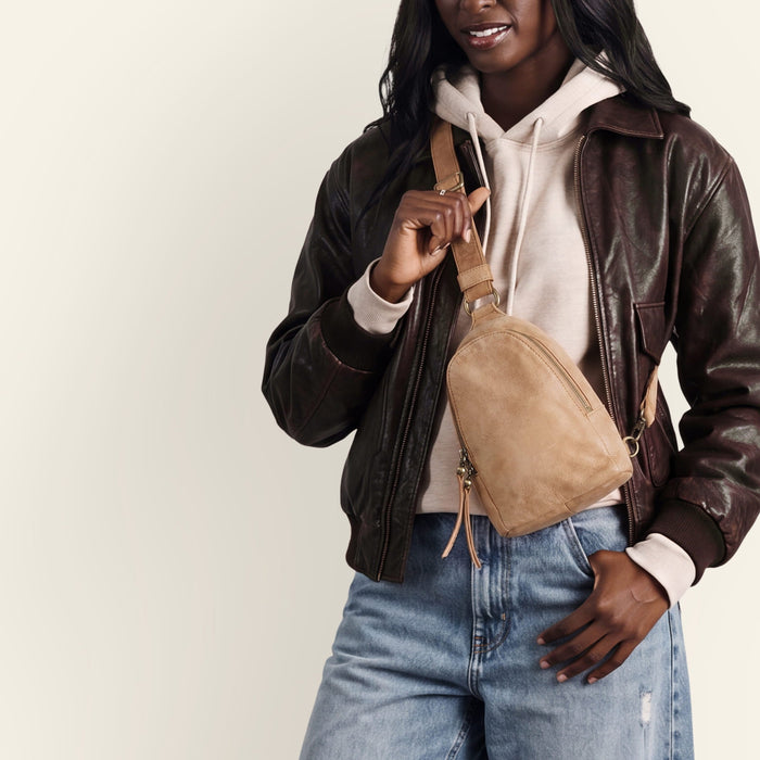A woman wearing a dark brown leather jacket, cream hoodie, and blue jeans carries a tan suede crossbody bag against a plain off-white background.