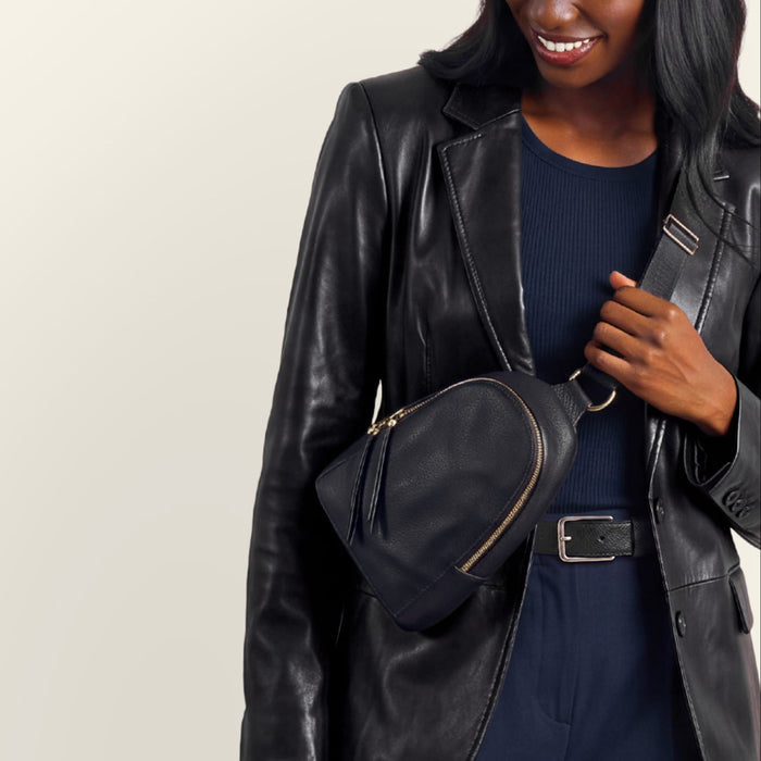 A woman wearing a black faux-leather blazer, navy blue top and pants, a brown and navy baseball cap, and carrying a matching black crossbody bag with gold-tone hardware, set against a plain light beige studio background.