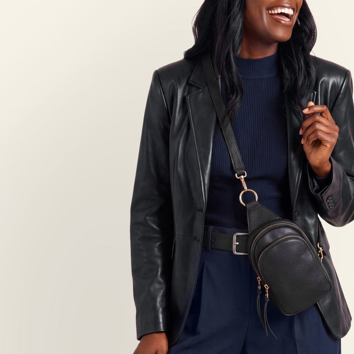A woman wearing a black leather blazer, navy blue ribbed top and trousers, a navy cap, and a black leather crossbody bag with gold-tone hardware, posing against a light beige studio background.