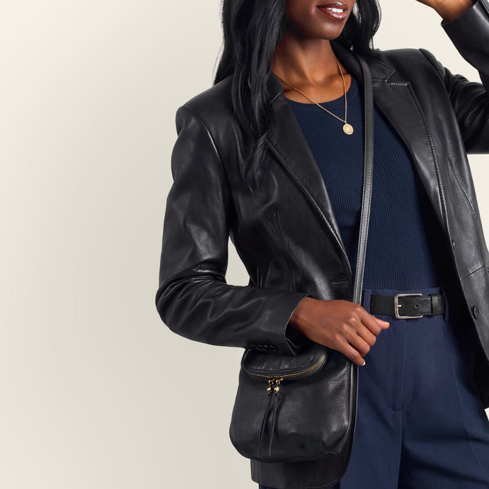 A model wearing a white square-neck t-shirt and blue jeans carries a black leather crossbody bag with gold-tone hardware and a tassel detail against a solid light gray background.