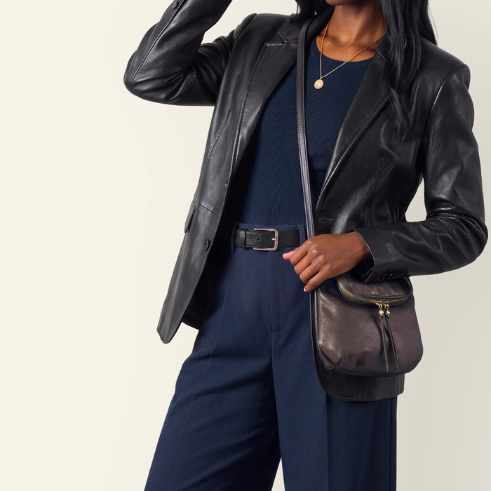 A model wearing a black leather blazer, navy blue ribbed top and trousers, and a dark brown leather crossbody bag with black trim and gold hardware, set against a plain light beige background.