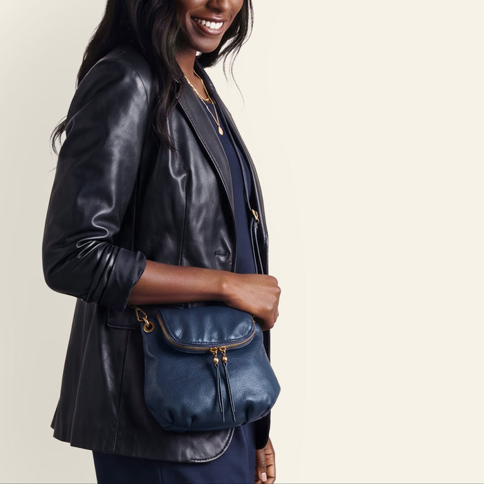 A woman wearing a black leather blazer and navy blue outfit carries a navy blue leather crossbody bag with gold-tone hardware against a light beige studio background.