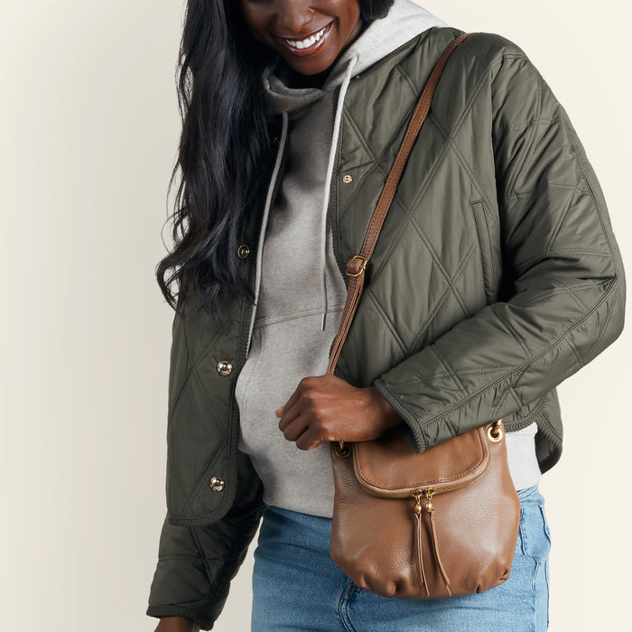 A woman wearing an olive green quilted jacket, a gray hoodie, and blue jeans carries a brown leather crossbody bag with gold-tone hardware against a plain light beige background.