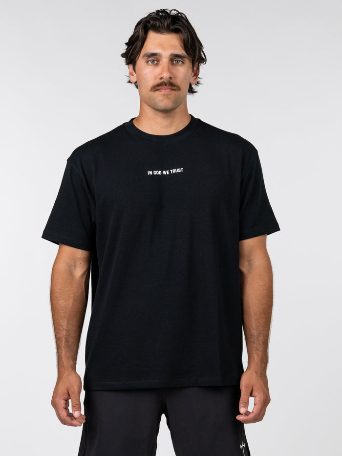 Man wearing a black God Bless America Premium Oversized Tee with "IN GOD WE TRUST" printed in white text on the chest, styled as Christian fitness apparel.