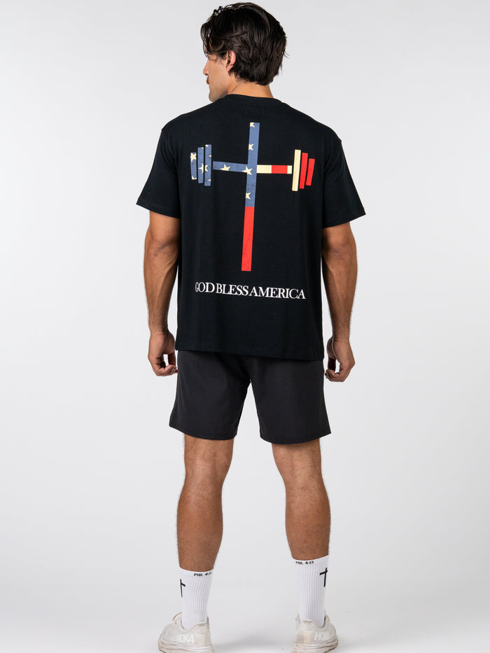Man wearing God Bless America Premium Oversized Tee with American flag-themed barbell cross graphic and text, paired with black shorts and white athletic socks.