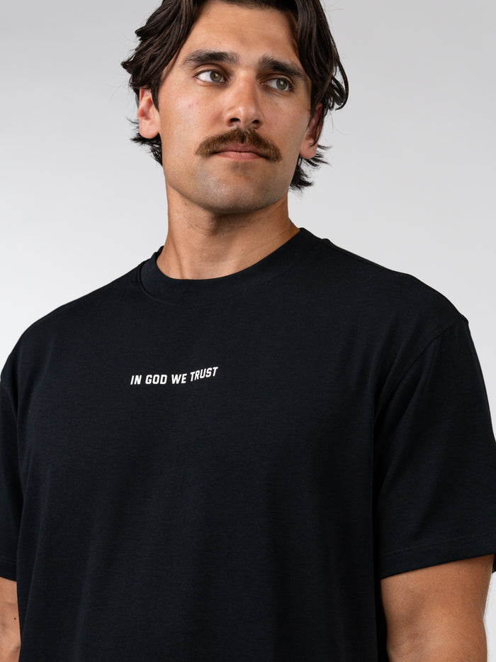 Man wearing a black God Bless America Premium Oversized Tee with the phrase "IN GOD WE TRUST" printed in white on the chest, showcasing faith-based casual apparel.