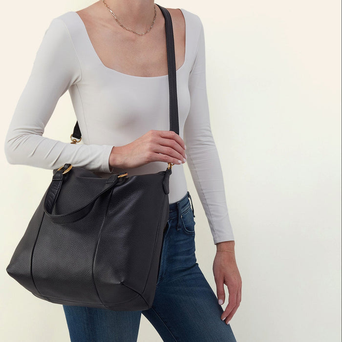 A woman wearing a white long-sleeve top and blue jeans carries a black leather shoulder bag with gold-tone hardware against a plain off-white background.