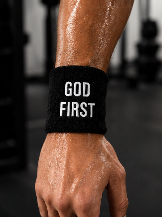 A close-up of a person's sweaty forearm and hand wearing a black wristband with the words 'GOD FIRST' in bold white capital letters, set against a blurred gym background.