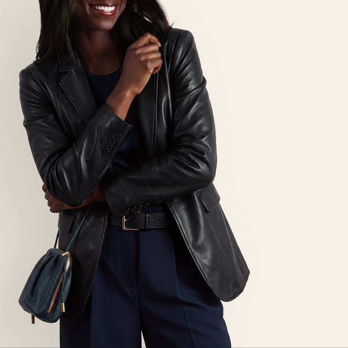 A woman wearing a black leather blazer over a navy top and navy trousers, holding a small navy handbag, against a plain light beige background.