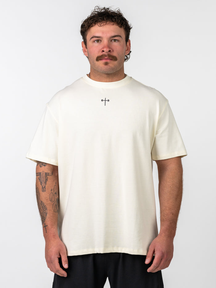 Man wearing a cream Isaiah 41:10 Premium Oversized Tee featuring a small black cross with barbell design on the chest, paired with black shorts.