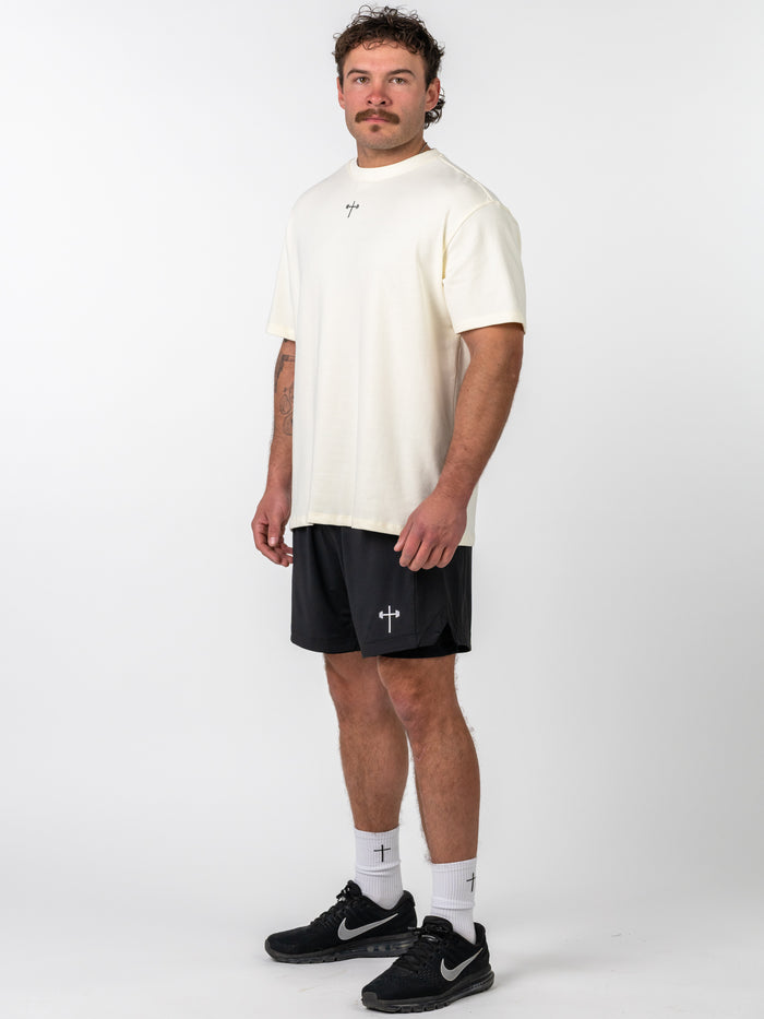 Man wearing the Isaiah 41:10 Premium Oversized Tee in cream with a small cross design on the chest, paired with black shorts and white socks featuring cross logos, standing against a plain background.