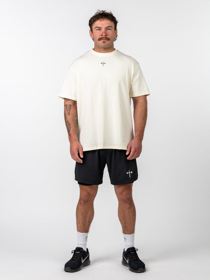 Man wearing a white Isaiah 41:10 Premium Oversized Tee with a small cross design on the chest, paired with black shorts and athletic shoes, standing against a white background.