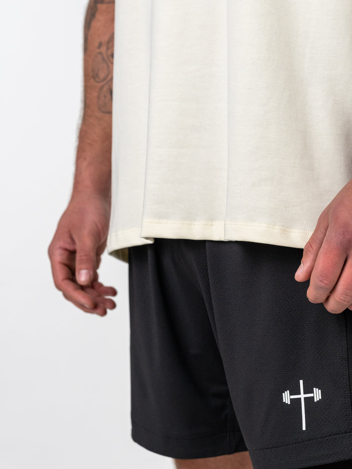 Close-up view of a person wearing the Isaiah 41:10 Premium Oversized Tee in light beige paired with black athletic shorts featuring a white barbell cross logo, representing Christian gym clothing.