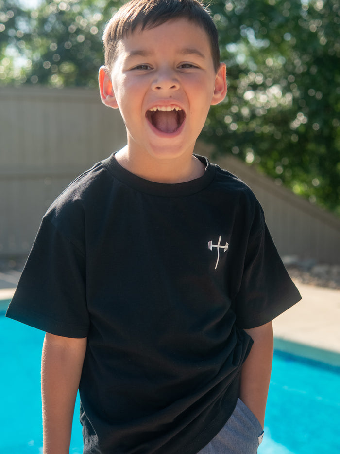 A young boy smiling and wearing a black Jesus is King Youth Tee featuring a white barbell cross logo on the front, standing outdoors by a pool in bright sunlight.