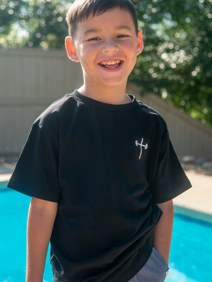 Young boy smiling outdoors wearing a black Jesus is King Youth Tee featuring a small white barbell cross logo on the front, styled as Christian gym clothing.