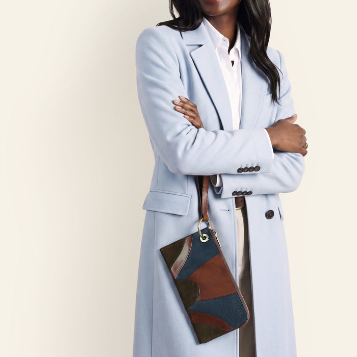 A woman wearing a light blue tailored coat over a white shirt, holding a multicolor patchwork clutch bag with a brown strap against a plain cream background.