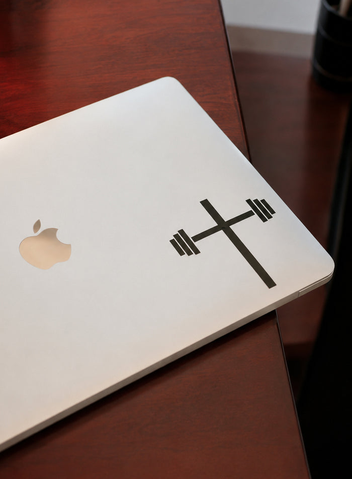 A silver laptop with a black decal featuring a cross combined with a barbell weight symbol, illustrating a faith and fitness theme from Laptop and Car Decals.