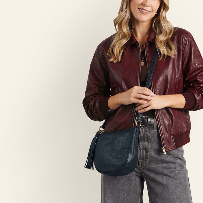 A woman wearing a burgundy leather bomber jacket and gray jeans, carrying a dark navy blue leather crossbody bag with gold hardware and a tassel.