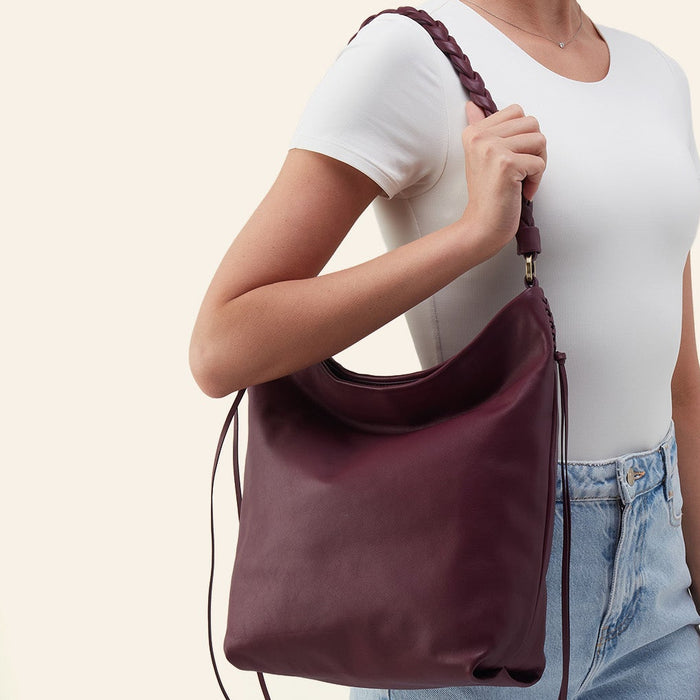 A woman wearing a white t-shirt and light blue jeans carries a dark burgundy leather slouchy shoulder bag with a braided strap against a light beige background.