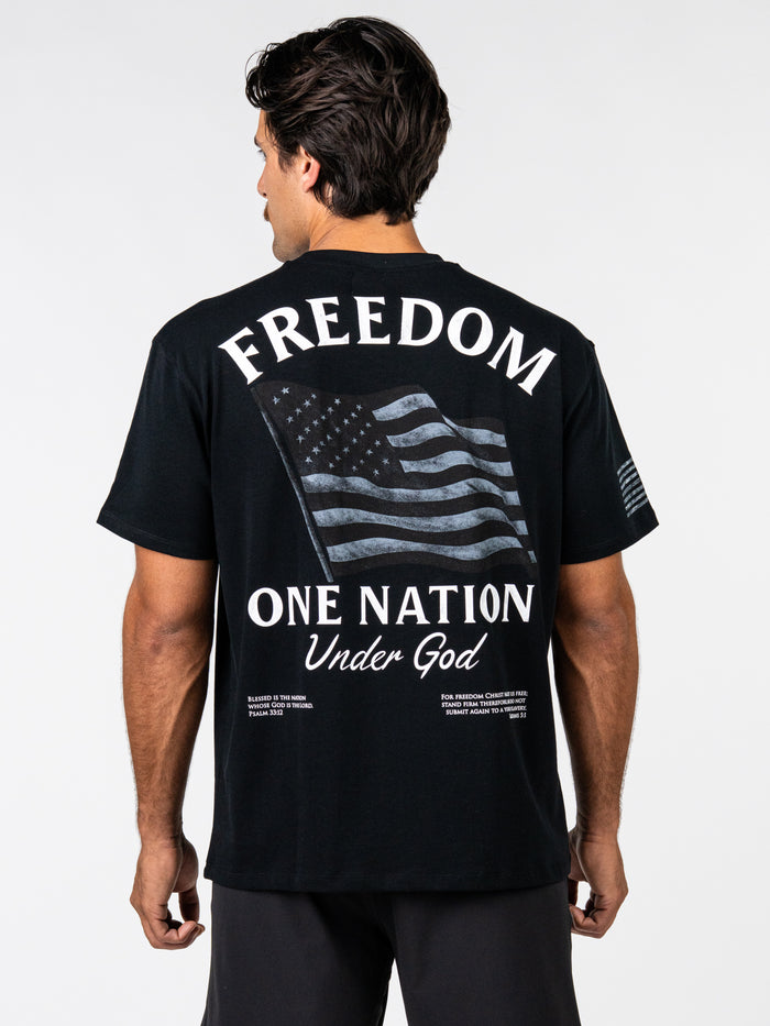 Man wearing a black One Nation Under God Oversized Tee featuring a distressed American flag design and the words "FREEDOM ONE NATION Under God" on the back, paired with black shorts.