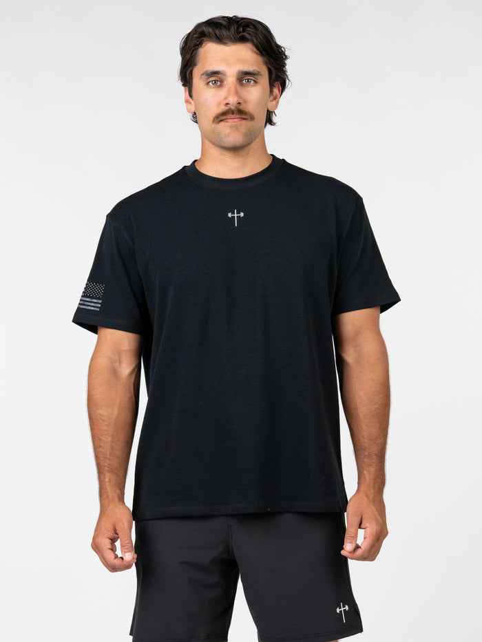 Man wearing a black One Nation Under God Oversized Tee featuring a small cross design on the chest and a subtle flag graphic on the sleeve, paired with black shorts.