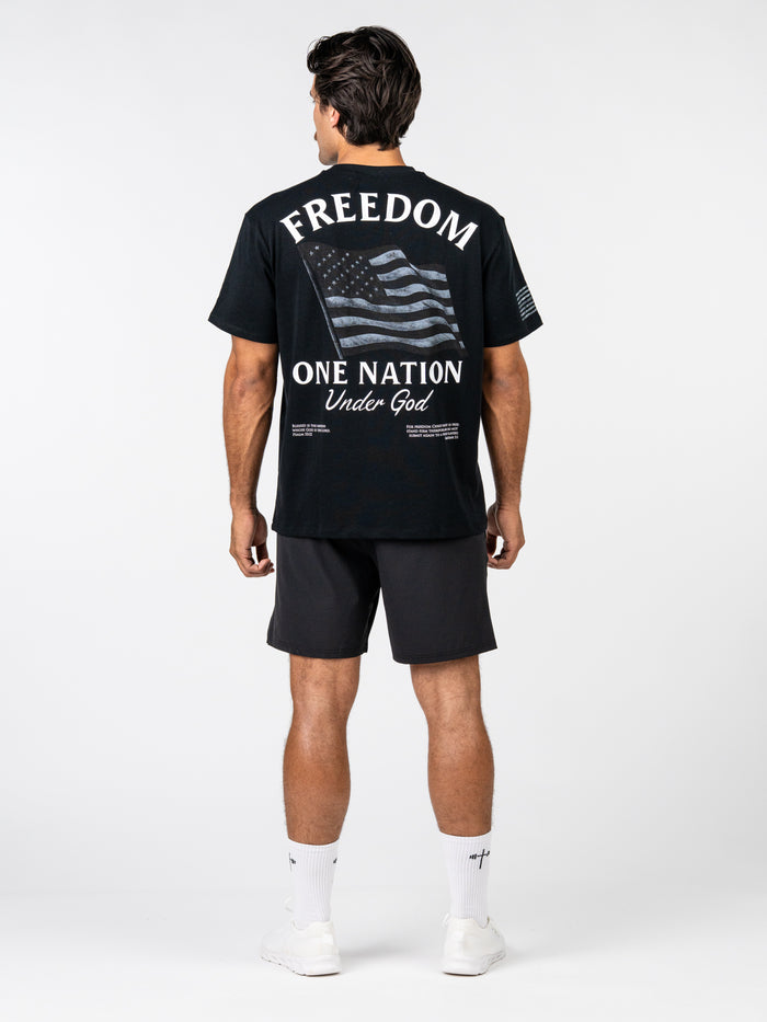 Man wearing the One Nation Under God Oversized Tee featuring a large American flag graphic and text promoting freedom, paired with black shorts and white athletic socks.