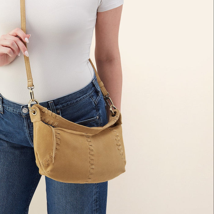 A woman wearing a white top and blue jeans carries a tan crossbody bag with a matching strap against a plain light beige background.