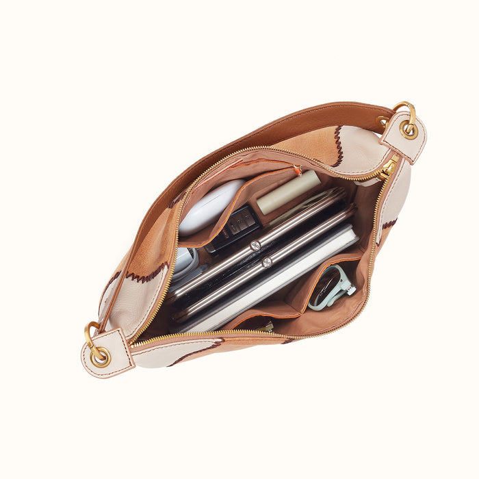 An open beige and brown leather shoulder bag with gold-tone hardware, shown from above to reveal its interior compartments filled with cosmetics and accessories, on a solid off-white background.