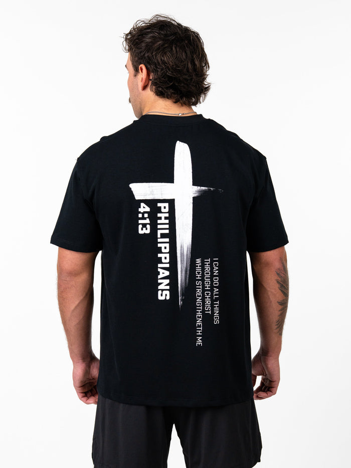 Man wearing a black Phil 4:13 Premium Oversized Tee featuring a large white cross design with the scripture reference Philippians 4:13 and the quote "I can do all things through Christ which strengtheneth me" on the back.