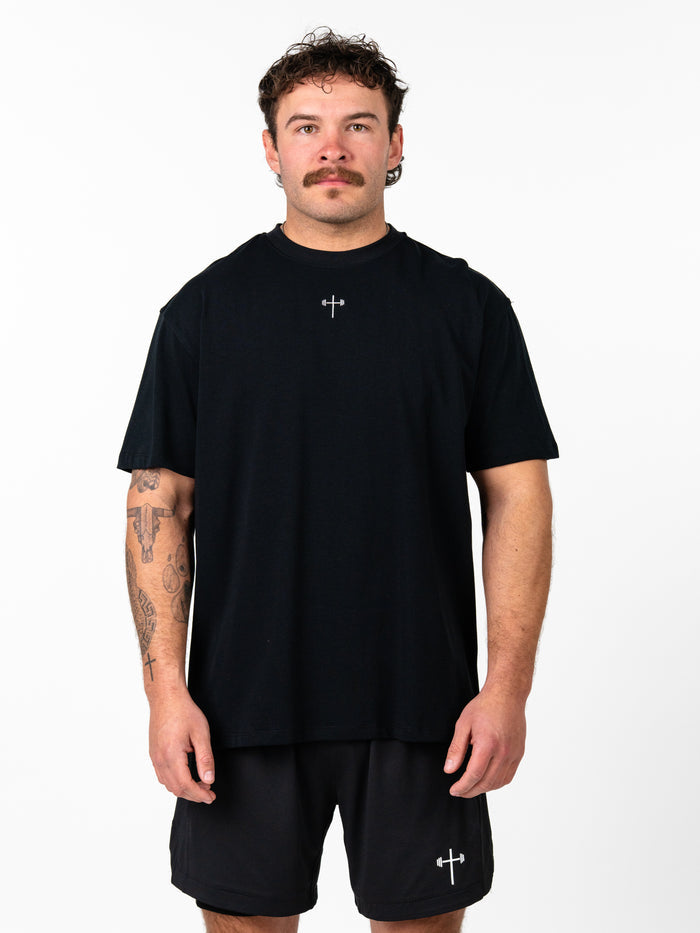 Man wearing a black Phil 4:13 Premium Oversized Tee featuring a small cross and barbell design at the chest, styled with matching black shorts for Christian fitness apparel.