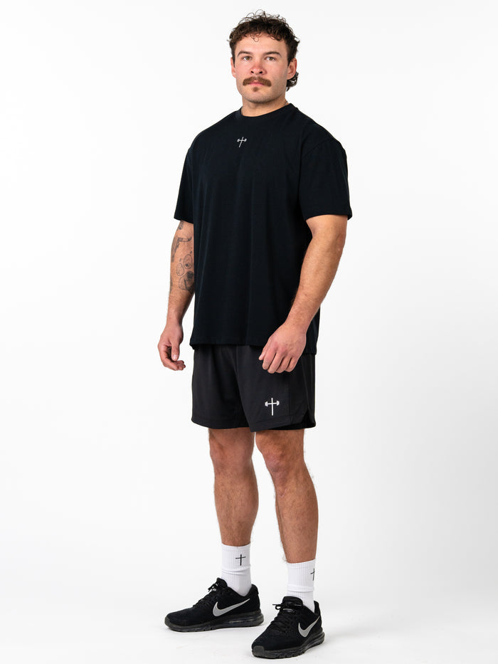 Man wearing black Phil 4:13 Premium Oversized Tee paired with black shorts featuring a Christian cross and barbell design, styled for faith-based fitness apparel.