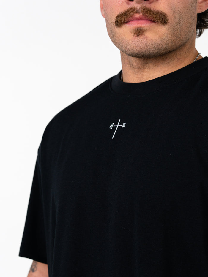 Close-up of a man wearing the Phil 4:13 Premium Oversized Tee in black, featuring a small white cross with a barbell design on the chest, representing Christian fitness apparel.