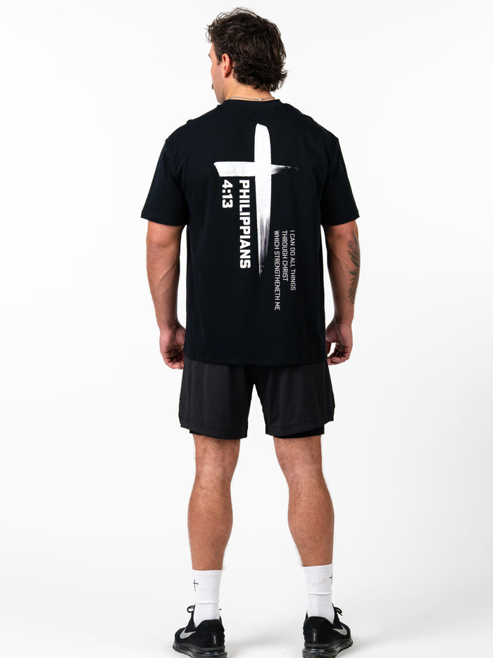 Man wearing black Phil 4:13 Premium Oversized Tee with white cross and scripture text printed on the back, paired with black shorts and white socks.