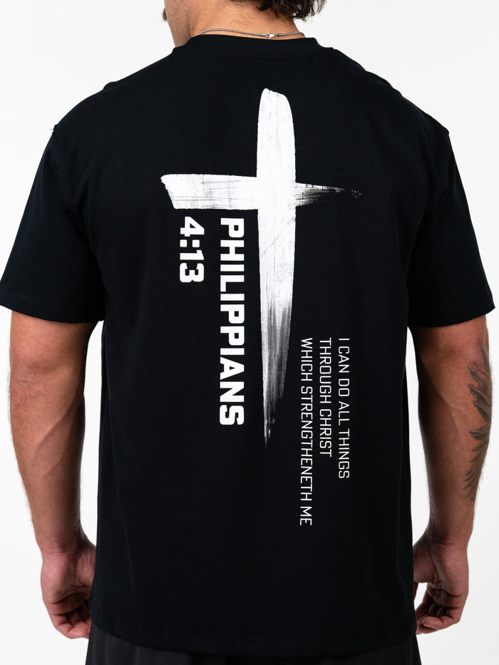 Man wearing a black Phil 4:13 Premium Oversized Tee with a large white cross design and the text "Philippians 4:13 I can do all things through Christ which strengtheneth me" on the back.