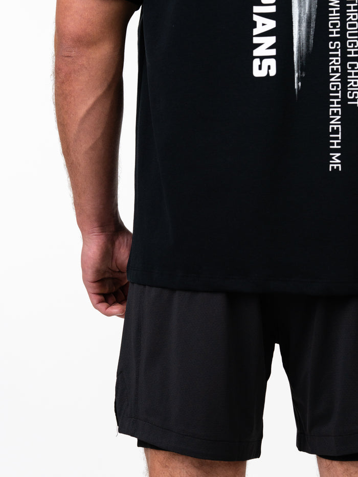 Close-up of a person wearing the Phil 4:13 Premium Oversized Tee in black paired with black athletic shorts, featuring vertical white text on the side related to Christian faith and strength.